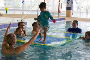 toddler smiling and running along mat in swimming lesson to parent's arms