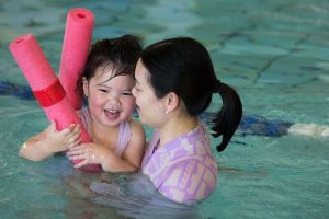 mother hugging and staring lovingly at daughter who is sitting on pool noodle in her lesson